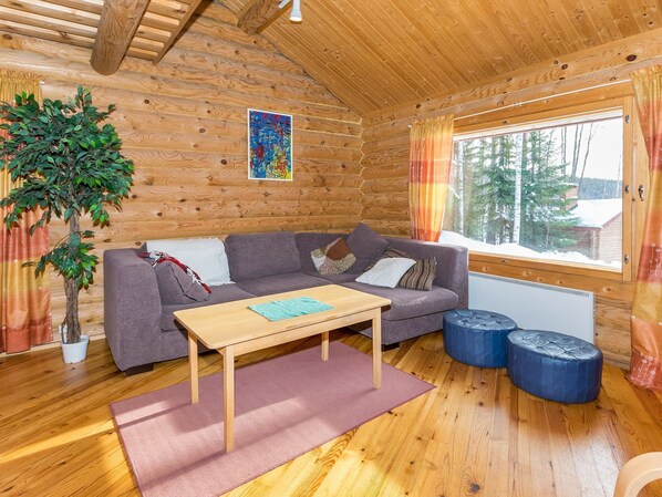 Indretning i en hyggelig hytte med væg af træstammer, en grå sofa, et træbord og et stort vindue med gardiner.