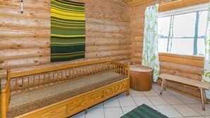 Interieur van een blokhut met een houten bank, een wandkleed met groene en gele strepen, een raam met gordijn en een houten kruk.