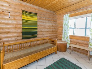 A log cabin interior with a wooden bench, a green and yellow striped wall hanging, a window with a curtain, and a wooden stool.