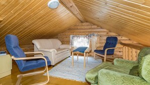 Een gezellige kamer onder de dakrand, met schuine wanden, houten lambrisering, een beige bank, blauwe en groene stoelen, een kleine tafel en een raam met gordijn.