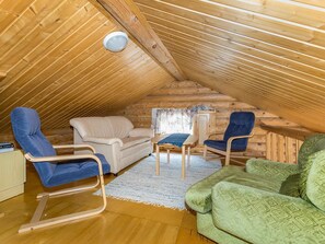 A cozy attic room with a sloped ceiling, wooden walls, a beige sofa, blue and green chairs, a small table, and a window with a curtain.