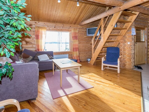 A cozy cabin interior with a wooden ceiling, a sofa, a chair, a small table, and a TV.