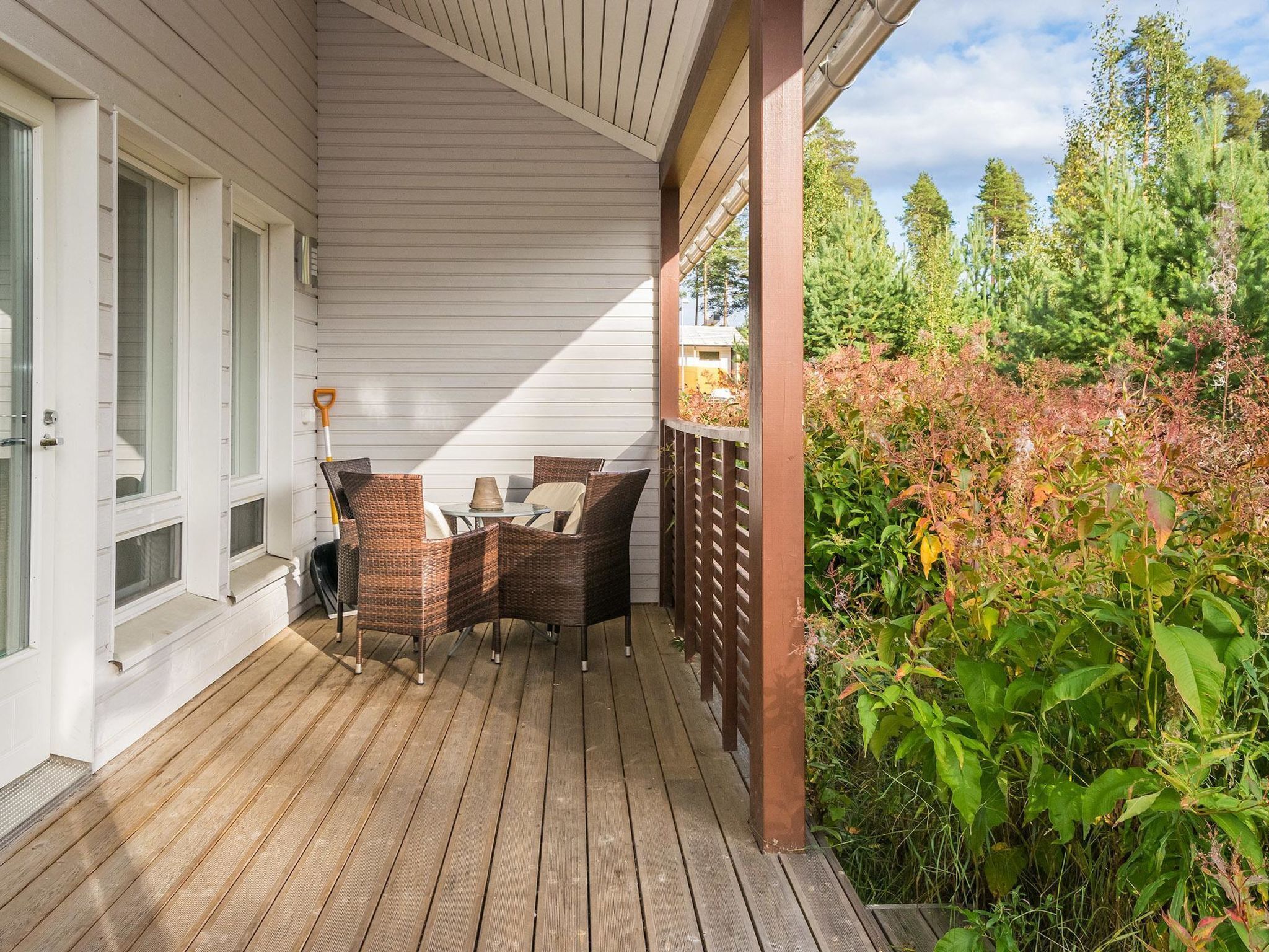 A wooden deck with wicker furniture and a glass table, surrounded by greenery and trees.