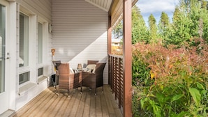 Terraza de madera con mobiliario de mimbre y una mesa de vidrio, rodeada de follaje y árboles.