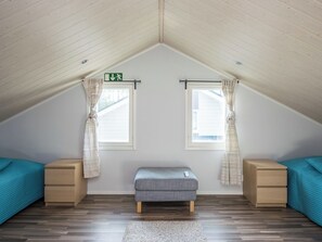 Una habitación con techo inclinado, dos ventanas con cortinas, un banco y mesitas de noche de madera.