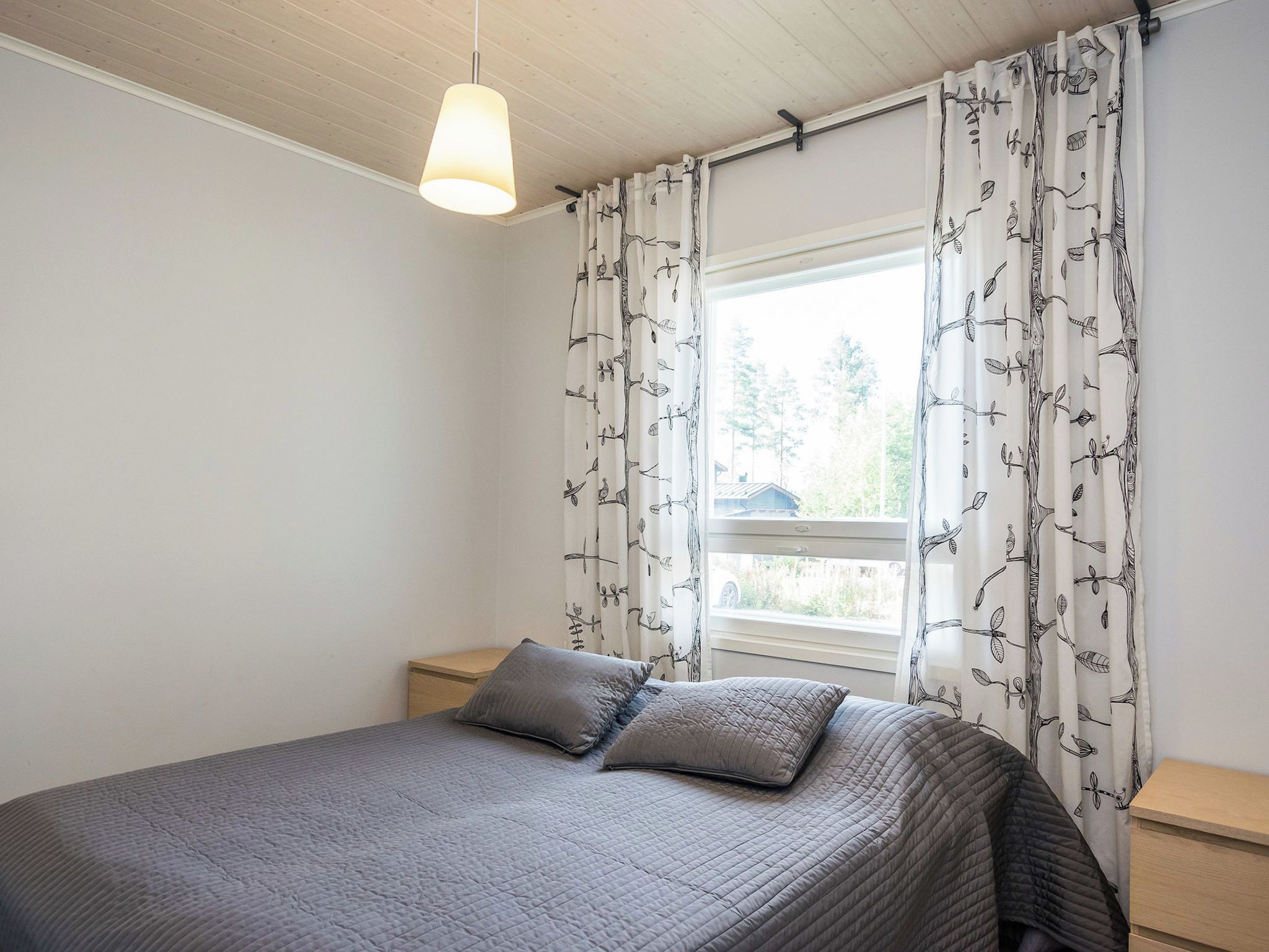 Une chambre à coucher avec un lit, deux oreillers, une table de chevet, une lampe suspendue et une fenêtre avec des rideaux.