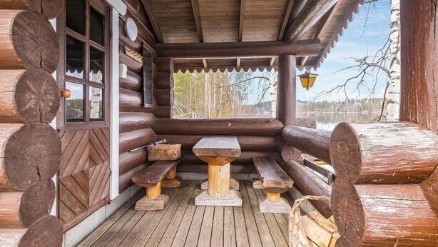 Una cabaña de troncos con terraza de madera, bancos y ventana con vistas a un bosque.