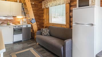 Una cocina compacta con estufa, horno y refrigerador, un sofá gris y una ventana con persianas.