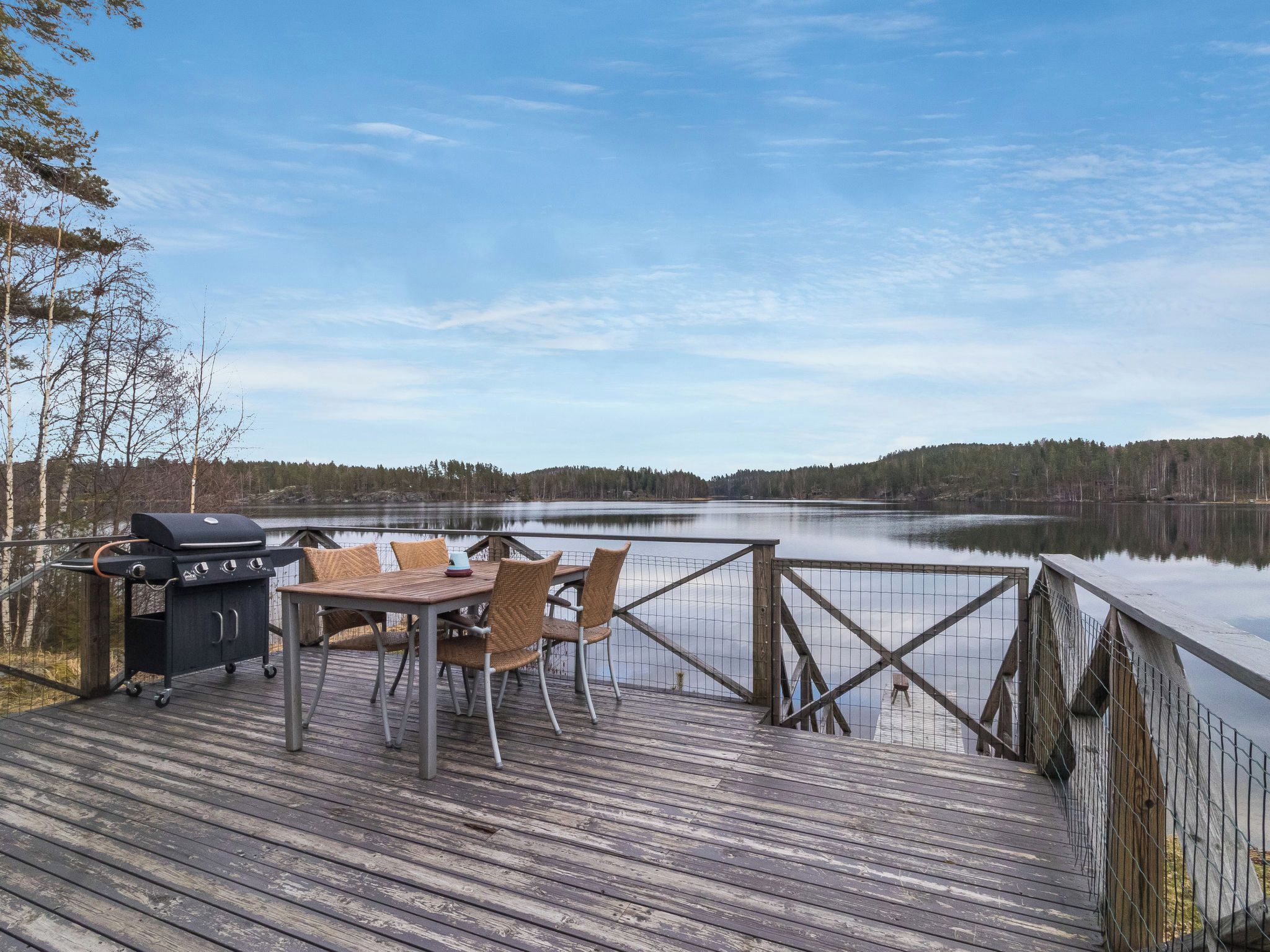 Een houten terras met een barbecue en tafel, met uitzicht op een kalm meer met een bebost oever.