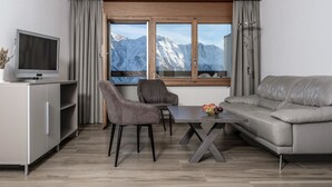 Een moderne woonkamer met een bank, twee stoelen, een eettafel met fruit, een televisie op een kast, en een raam met uitzicht op een berg.