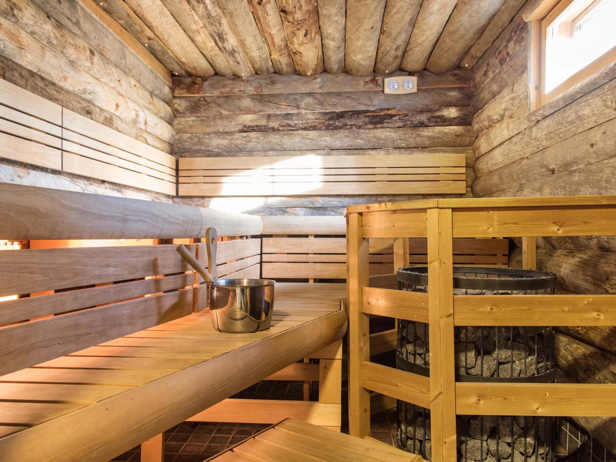 A sauna room with wooden benches, a metal bucket, and a stone fire pit.