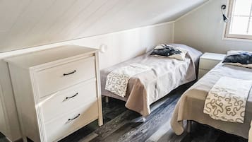 Een kleine, eenvoudige kamer met een schuine kap, een witte dressoir met lades, een bed met een bedovertrek met dessin en een raam met zonwering.