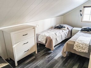 Een kleine, eenvoudige kamer met een schuine kap, een witte dressoir met lades, een bed met een bedovertrek met dessin en een raam met zonwering.