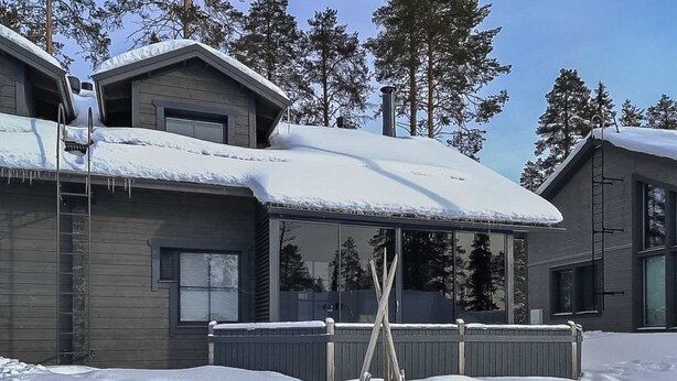 Una casa moderna con un amplio ventanal, una terraza de madera y un techo cubierto de nieve.