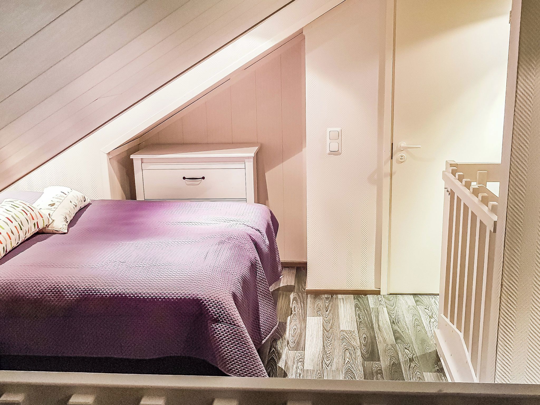 A bedroom with a sloped ceiling, a bed with a purple bedspread, a white nightstand, and a door with a white frame.