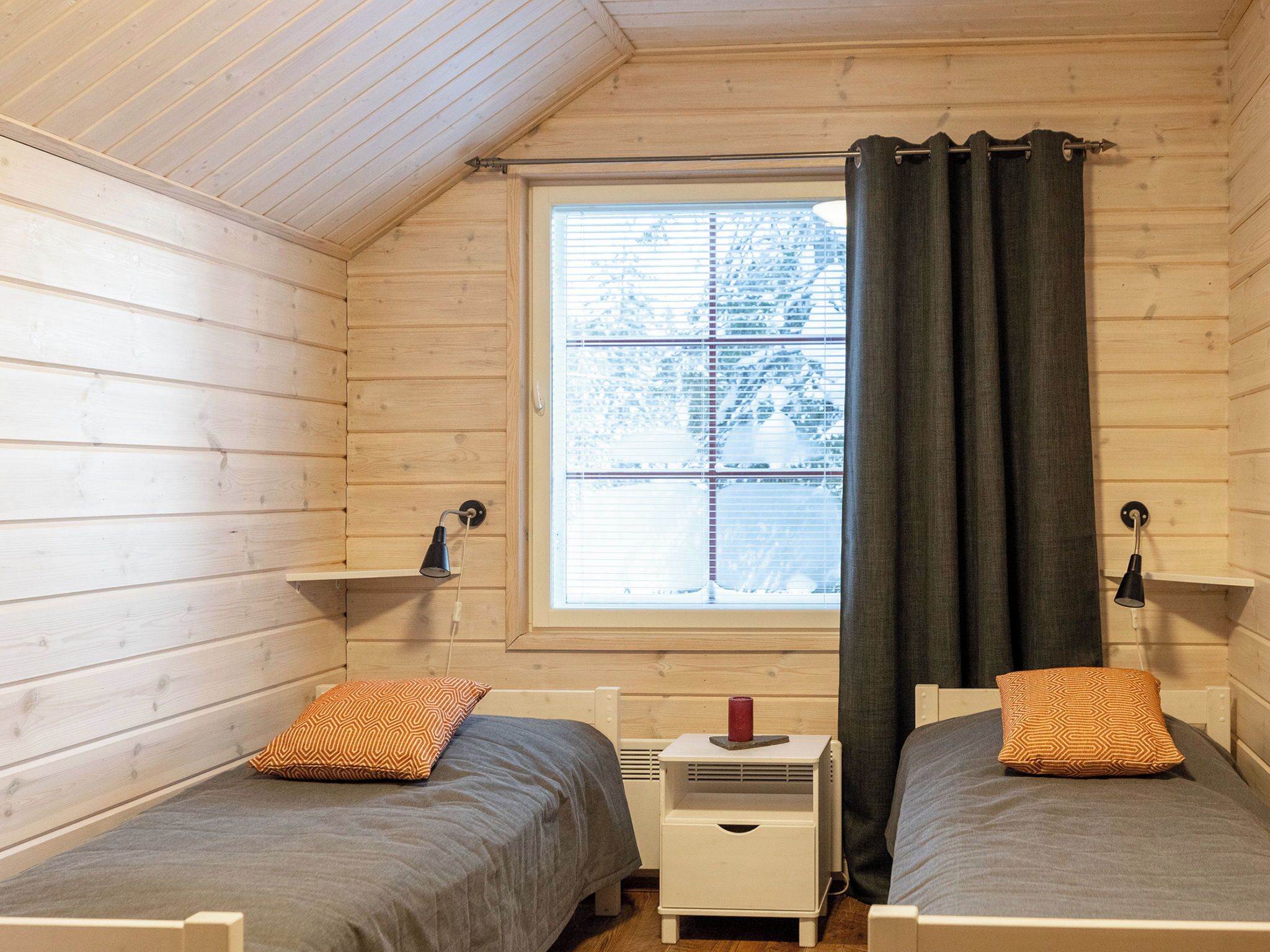 A small, cozy room with two single beds, a window with blinds, a heater, and a small table with a lamp.