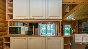 Interior de una cabaña de madera con zona de cocina que incluye armarios blancos, un microondas y una mesa de madera.