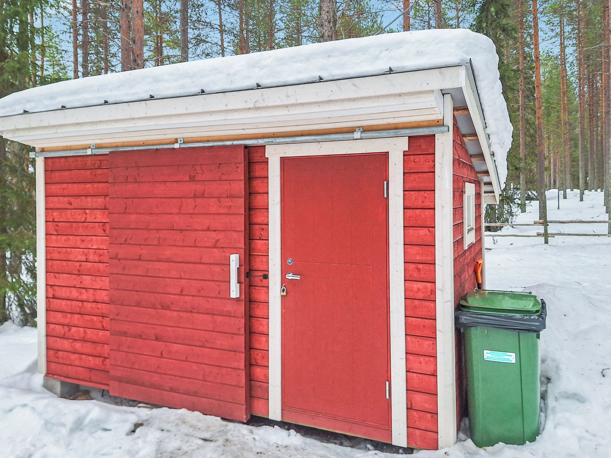 Punainen puinen varasto, jossa on valkoinen ovi ja sen vieressä vihreä roskakori. Varaston ympärillä on lunta ja puita.