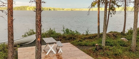 Une terrasse en bois avec un hamac et deux chaises blanches, donnant sur un lac paisible bordé d’arbres.