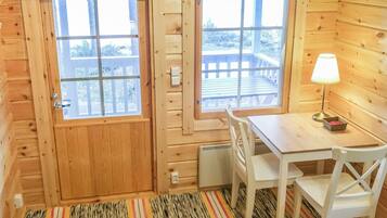 A wooden cabin interior with a dining area, two windows, a table, chairs, a lamp, and a rug.