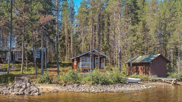 Een chalet met een veranda en een tuinhuisje aan een meer, omringd door bomen.