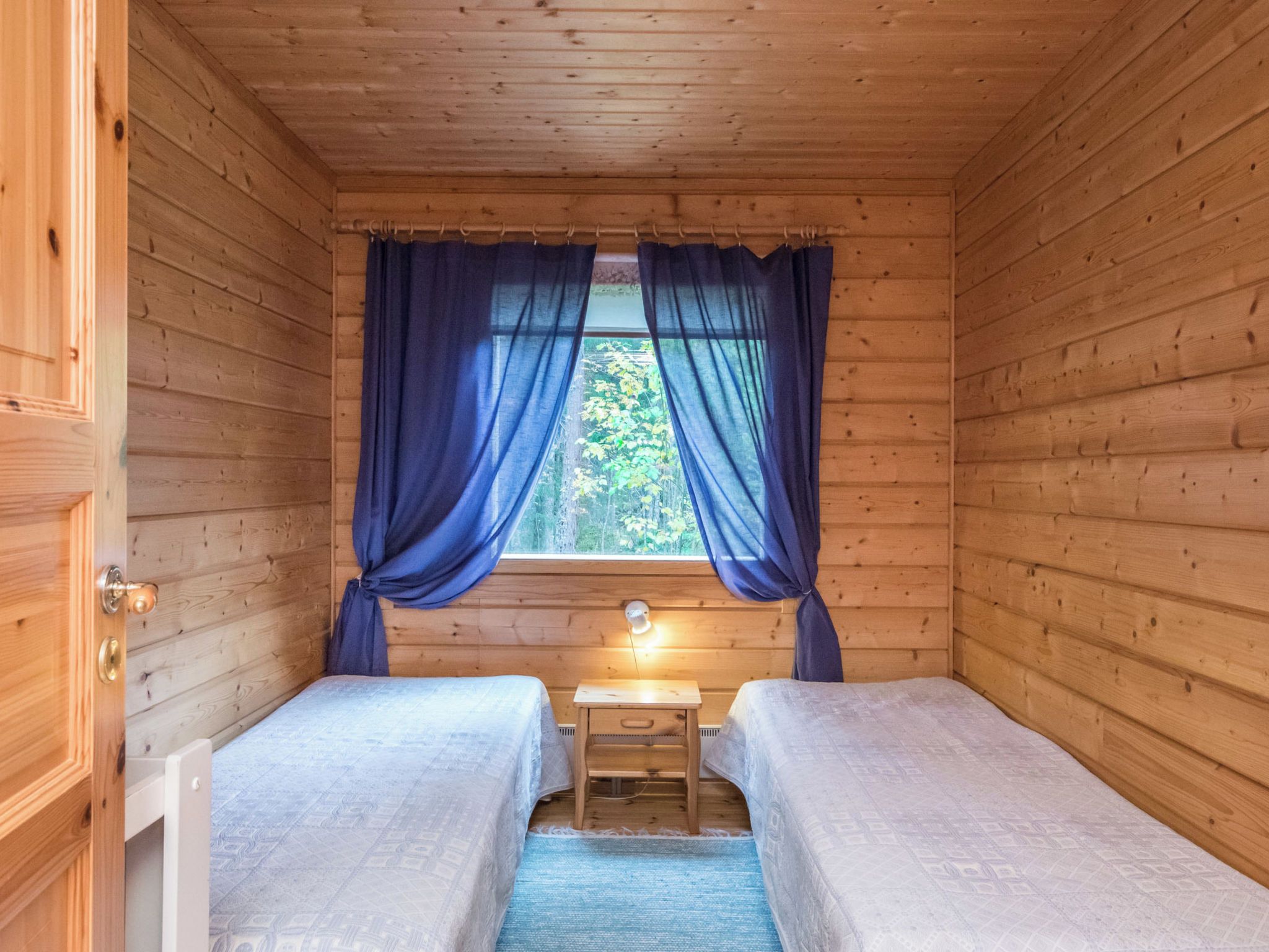 Eine kleine Holzhütte mit zwei Betten, einem Fenster mit Vorhängen, einem Nachttisch und einer Lampe.