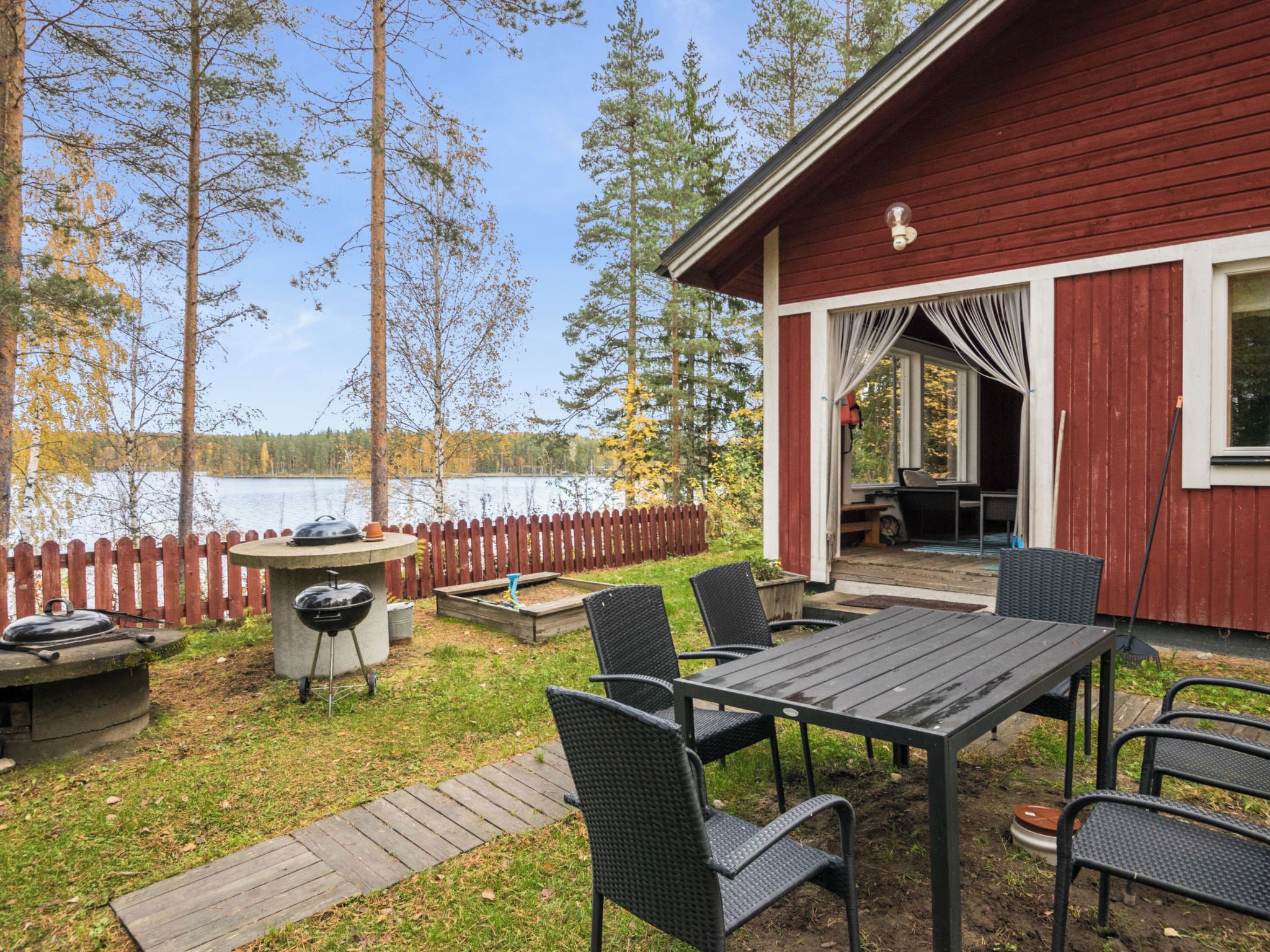 Eine Holzterrasse mit Grill und Gartenmöbeln, mit Blick auf einen See und Bäume im Hintergrund.