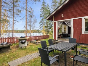 Een houten terras met een barbecue en tuinmeubilair, met uitzicht op een meer met bomen.
