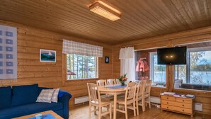 A cozy cabin interior with a wooden floor, a dining table with chairs, a blue sofa, a TV mounted on the wall, and a window with a view of trees and a lake. - Aamuntorkku by Interhome (Punkaharju)