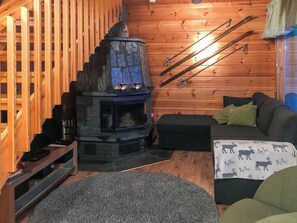 Interior acolhedor de uma cabana com lareira de pedra, paredes de madeira, um sofá e esquis encostados na parede.