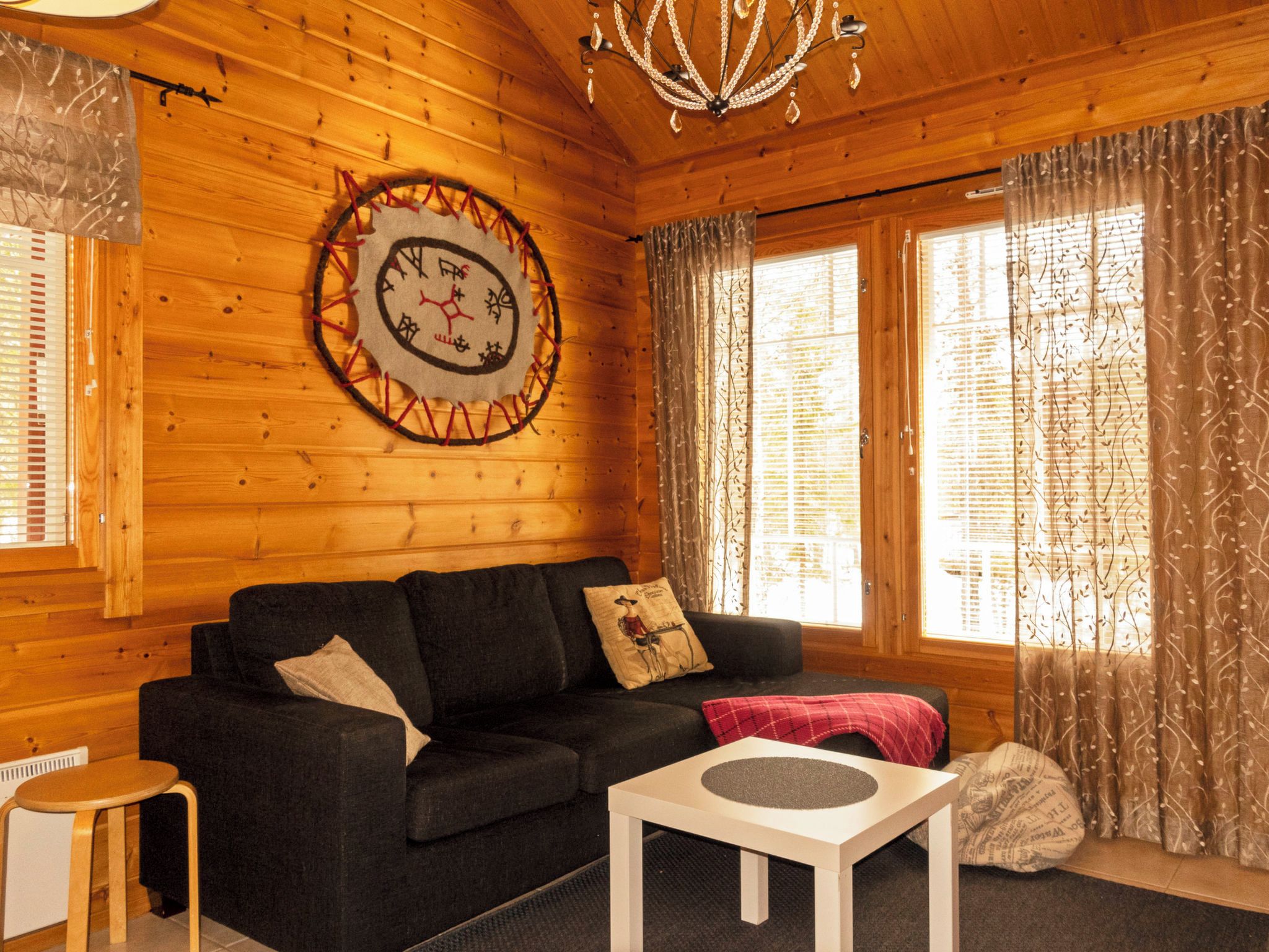 A cozy cabin interior with a wooden wall, a round wall clock, a sofa, a small table, and a window with curtains.