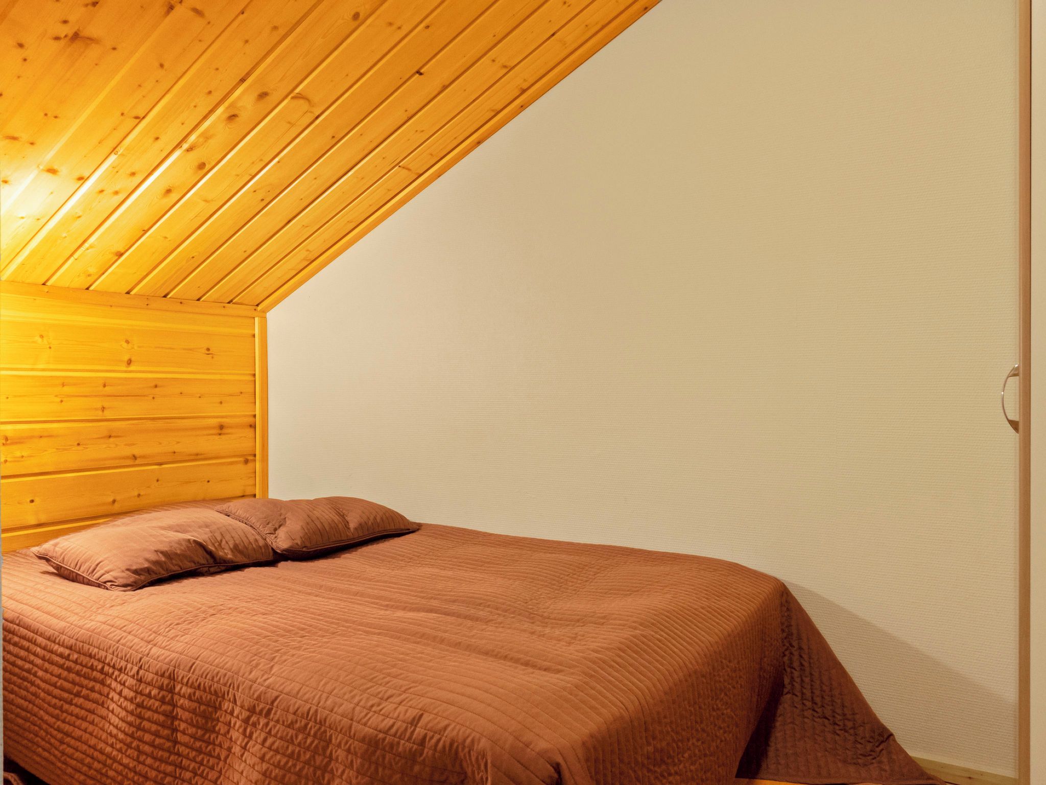 Een eenpersoonsbed met een bruin sprei in een kamer met houten balken en witte muren.