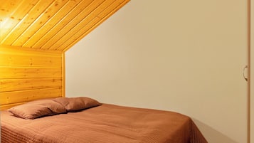Una cama individual con colcha marrón en una habitación con vigas de madera en el techo y paredes blancas.