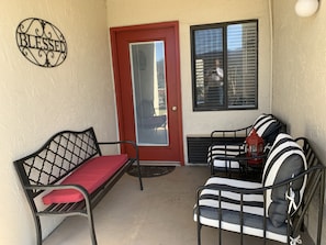 A porch with a red door, a metal bench with a red cushion, and a metal chair with striped cushions.