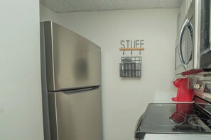 Una cocina compacta con un refrigerador de acero inoxidable, una cafetera roja y un estante de almacenamiento instalado en la pared con la etiqueta 'STUFF'.