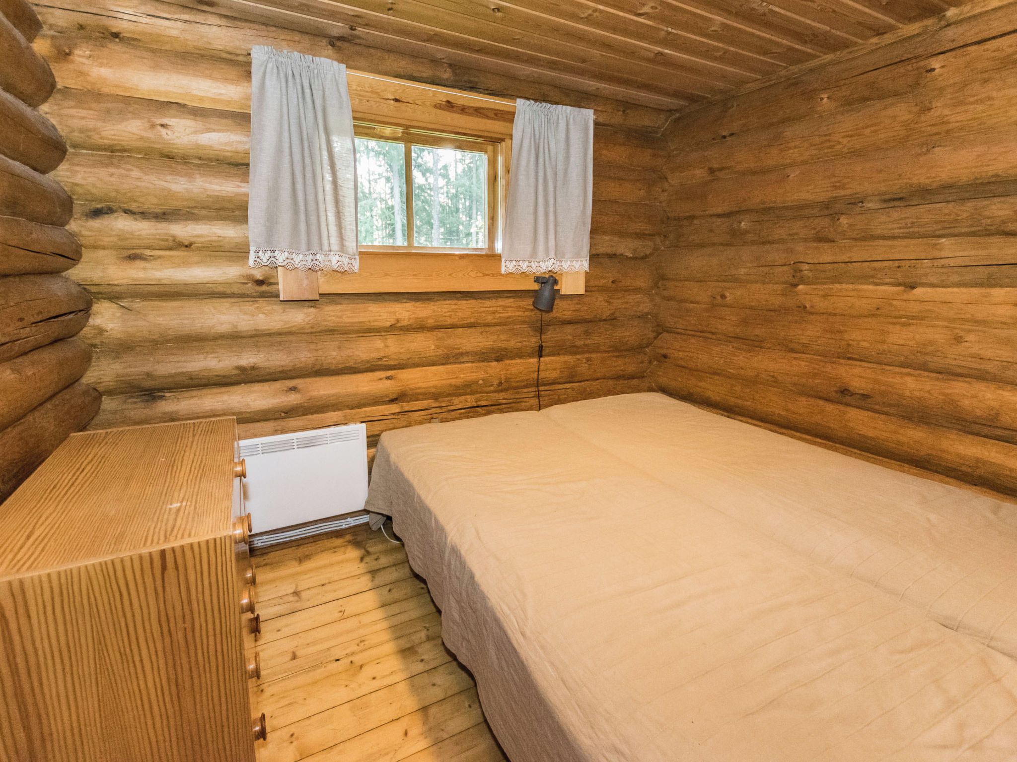 Une chambre de style rustique, aménagée dans une cabane en rondins, avec un lit simple, une petite table et une fenêtre avec des rideaux.