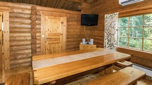 A wooden cabin interior with a large dining table and benches, a television mounted on the wall, and a window with a view of greenery.