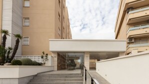A modern building with a flat facade, a staircase leading to an entrance, and a small garden area with palm trees. - Front Beach by Interhome (Calafell)
