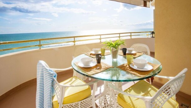 A round glass table with white chairs and yellow cushions on a balcony overlooking the sea. - Front Beach by Interhome (Calafell)