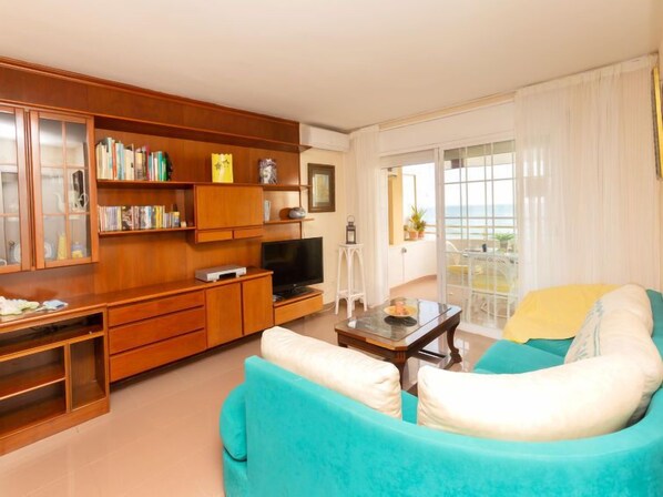 A living room with a sofa, TV, wooden shelves, and a balcony with curtains. - Front Beach by Interhome (Calafell)
