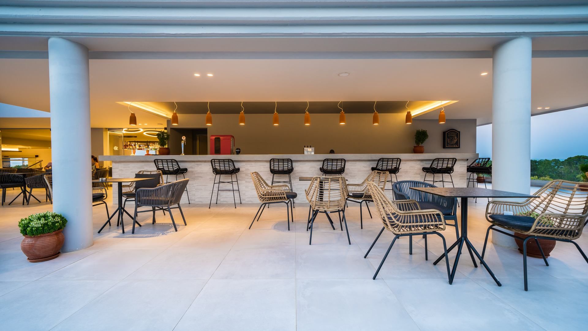 Un área de comedor al aire libre moderna con sillas y mesas de mimbre, una barra y luces colgantes.