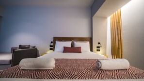 Quarto de hotel moderno com uma cama bem arrumada, uma mesinha de cabeceira com abajur e um espaço de estar com uma poltrona e uma mesa pequena.