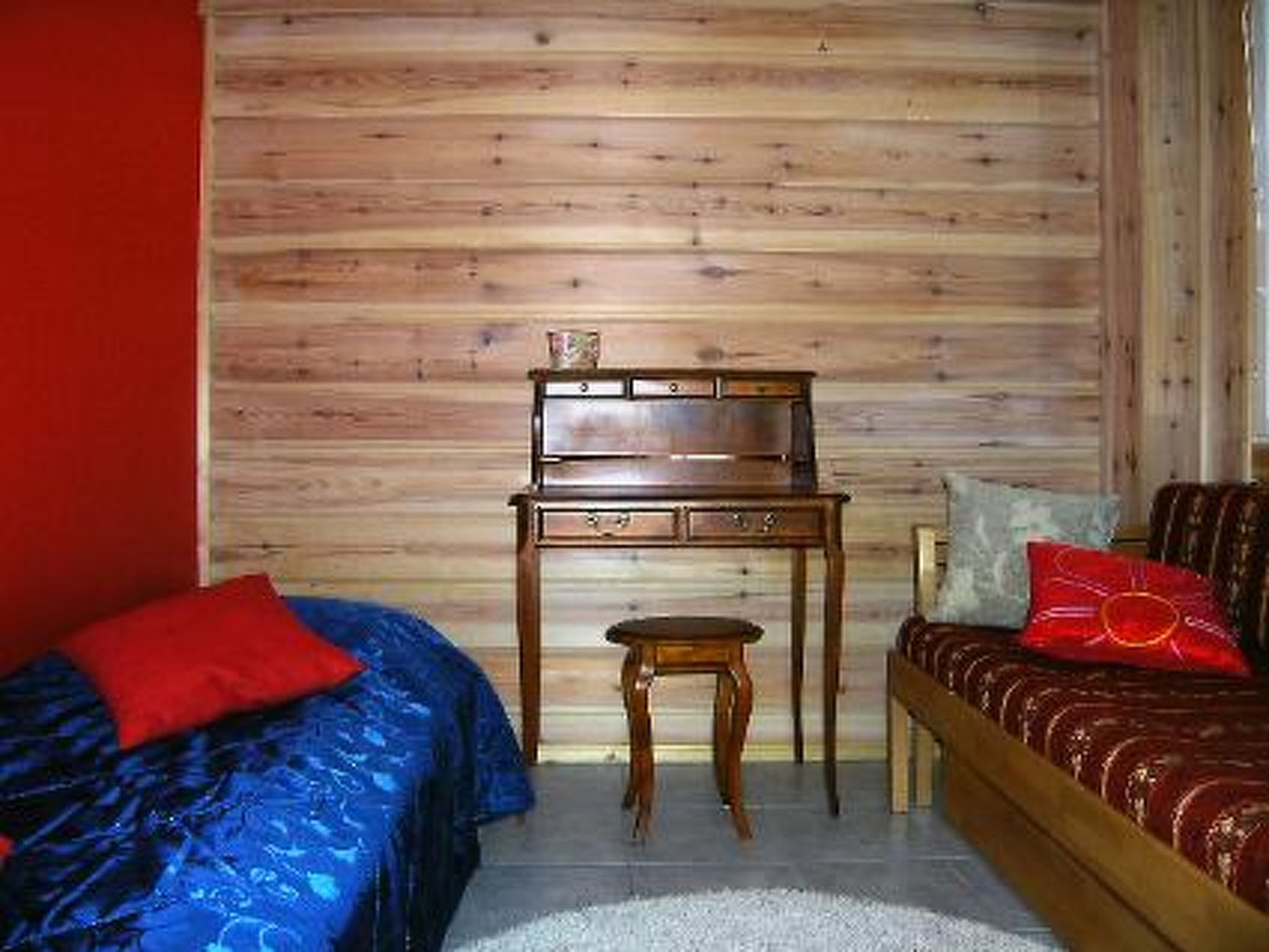 Een kamer met een houten wand, een bed met een rood kussen, een houten nachtkastje en een stoel met een rood kussentje.