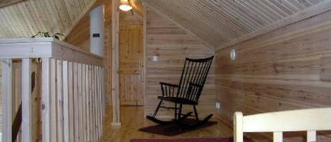 Un dormitorio estilo altillo acogedor, con techo de madera, una mecedora, una cama con colcha y una pequeña mesa con silla.