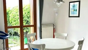 A dining area with a round white table and four white chairs with blue cushions. There is a pendant light above the table, a radiator on the wall, and a framed picture.