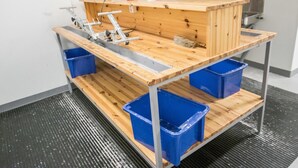 A wooden workbench with blue bins, a sink, and a metal stool.
