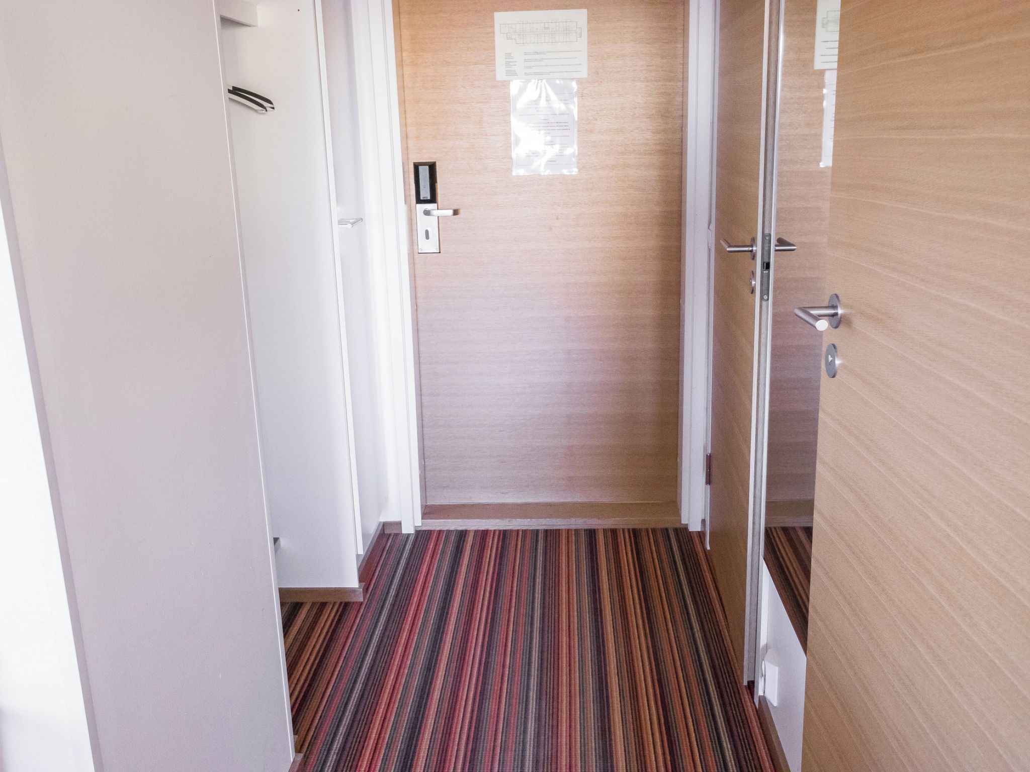 A hallway with a striped carpet, a wooden door, and a white wall with a notice.