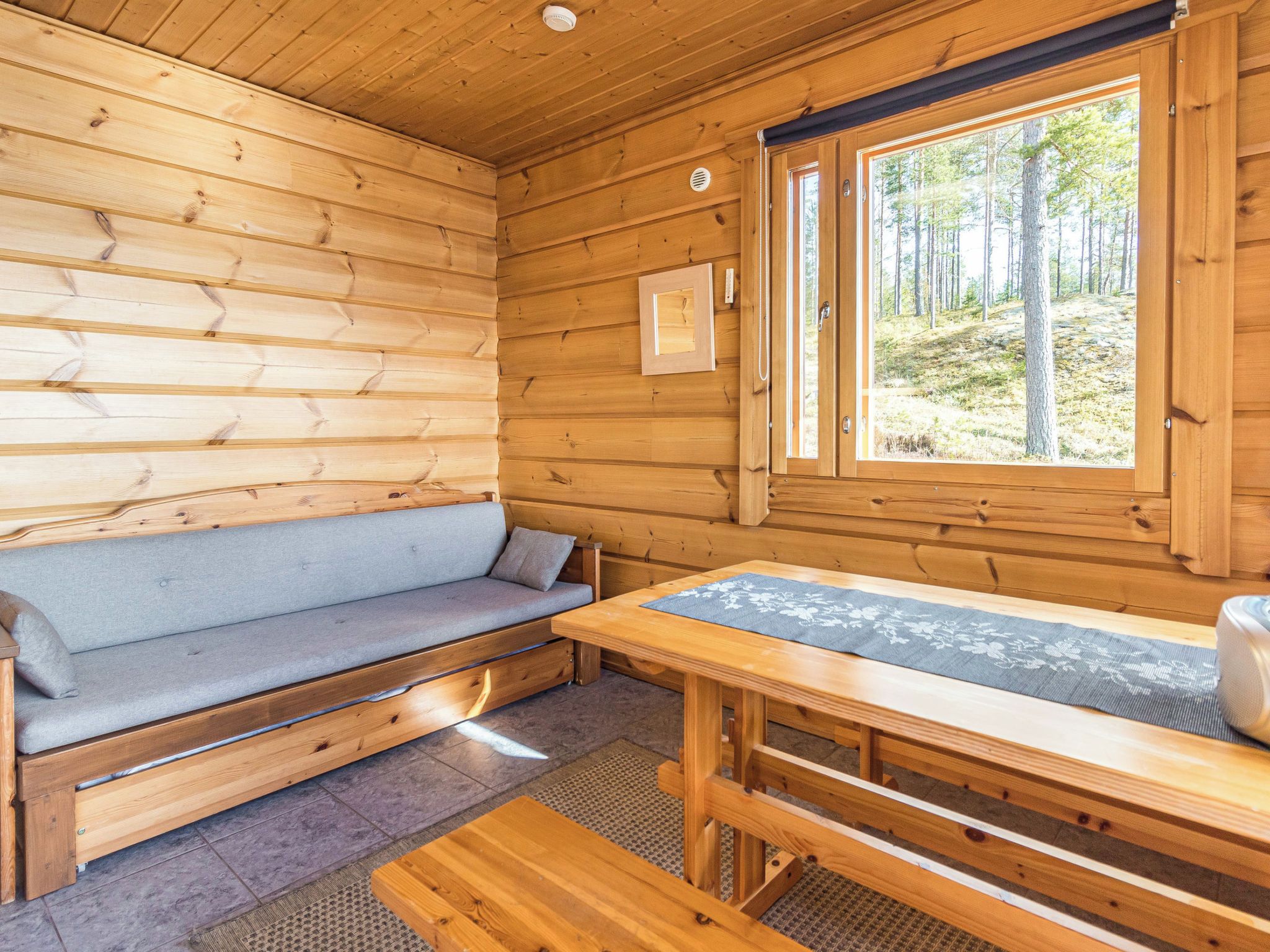 Interior de una cabaña con un banco de madera, una mesa y una ventana que da a un bosque.