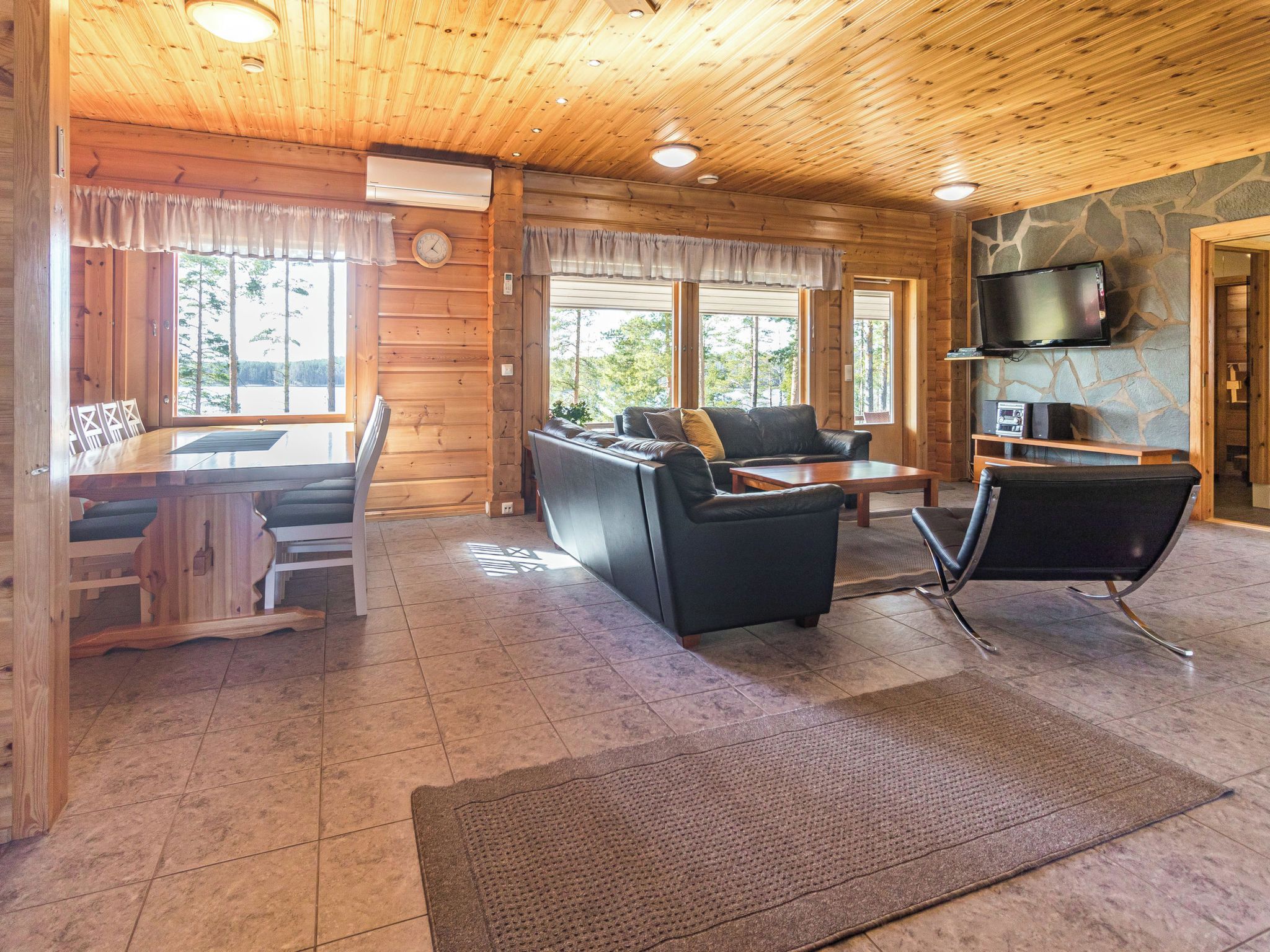 L’intérieur d’une cabane en rondins comprend un coin repas, un salon avec un canapé et des fauteuils, un téléviseur fixé à un mur en pierre et une cheminée.