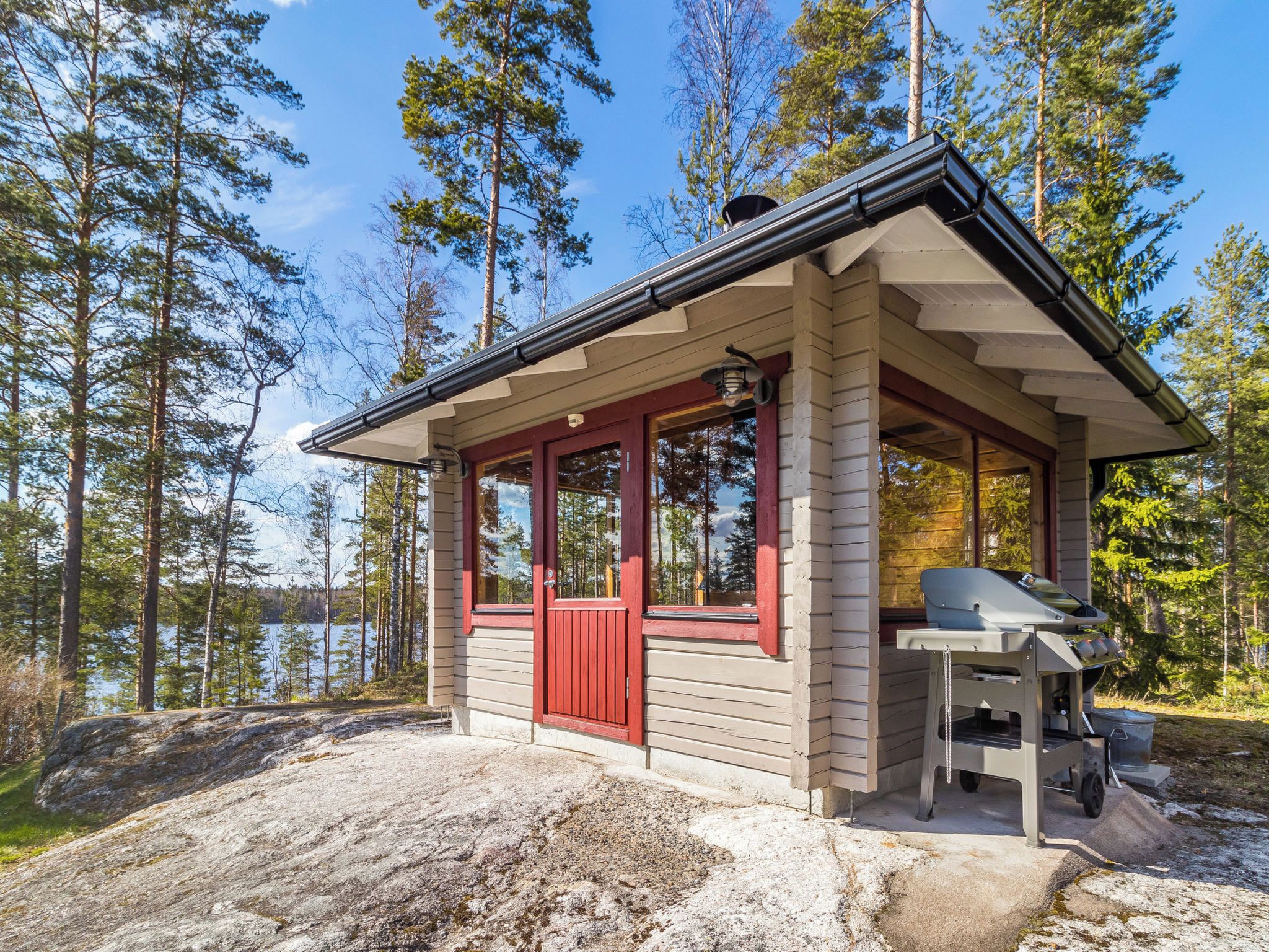Pieni mökki, jossa on punainen ovi, katettu kuisti ja ulkogrilli.